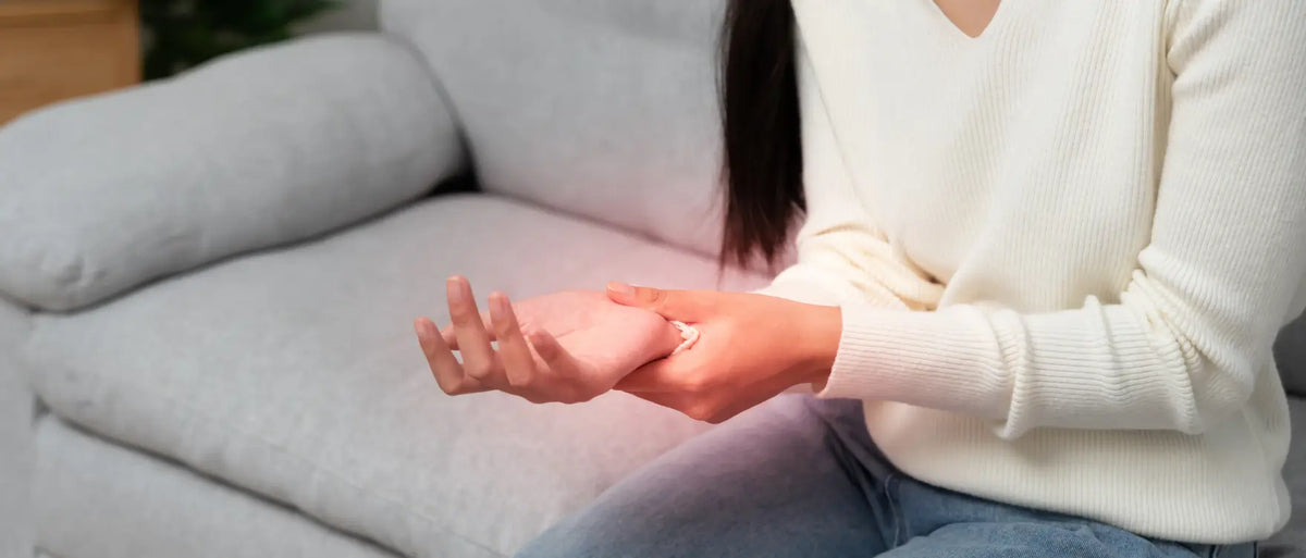 Woman in a cream sweater sitting on a light gray sofa, gently holding her wrist, representing everyday joint discomfort that can follow you into restless nights