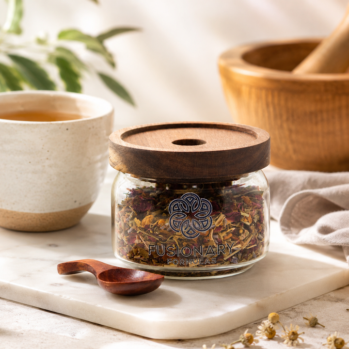 Glass jar with wooden lid containing dried herbs, placed on a marble surface with a cup of tea and wooden spoon in the background.